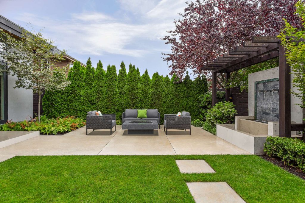 Porcelain Pavers - Swick's Landscaping Ltd. Modern backyard seating area with gray wicker furniture and a central fire table on a tiled patio, surrounded by tall evergreen hedges.