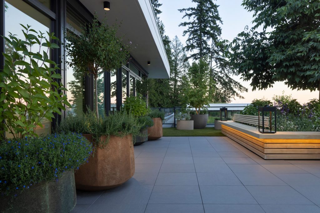 Swicks_3101_Burfield_Web-14-7060 - Swick's Landscaping Ltd. Terrace with tall potted plants along a glass-walled building, wooden bench seating, and trees in the background at dusk.