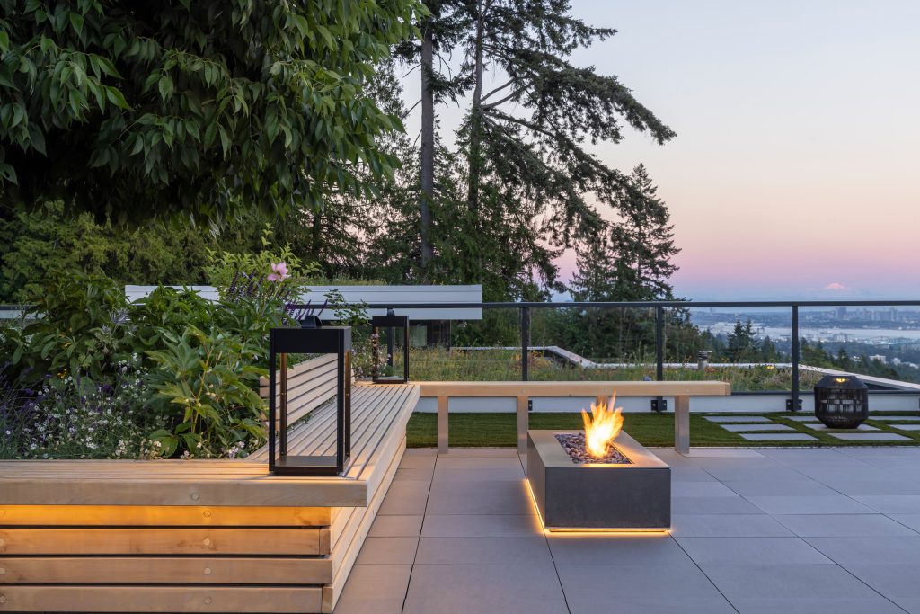 Swicks_3101_Burfield_Web-15-7079 - Swick's Landscaping Ltd. Modern rooftop deck at dusk with wooden steps, lush planters, and a glowing rectangular fire pit against a city skyline.