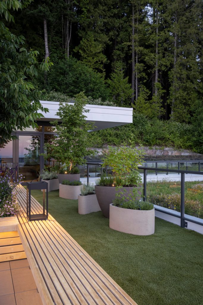 Swicks_3101_Burfield_Web-17-7084 - Swick's Landscaping Ltd. Modern terrace garden with large potted plants and a wooden slatted bench alongside a glass railing, against a forest backdrop