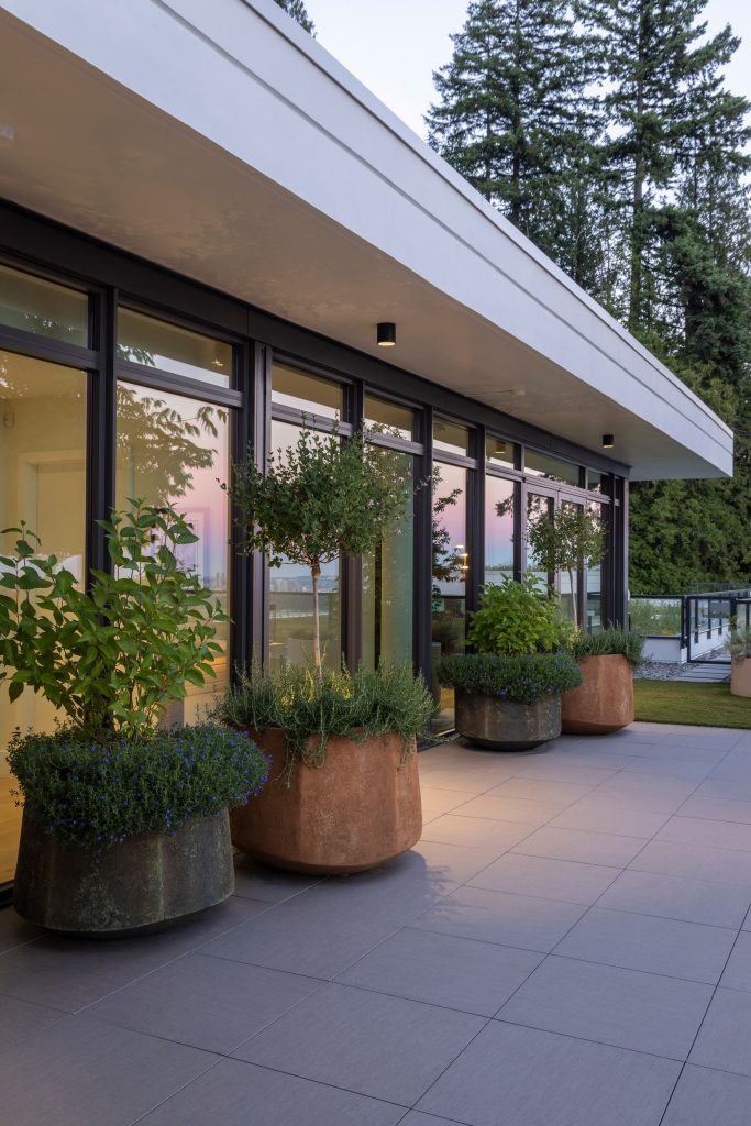 Swicks_3101_Burfield_Web-18-7086 - Swick's Landscaping Ltd. Modern glass-walled building on a tiled terrace with large rust-colored planters and green shrubs, reflecting a pink sunset in the windows.