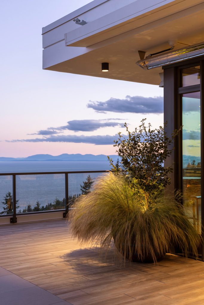 Swicks_3101_Burfield_Web-21-7110 - Swick's Landscaping Ltd. Balcony with wooden deck, ornamental grasses in a planter, and a glass railing overlooking the ocean at sunset.