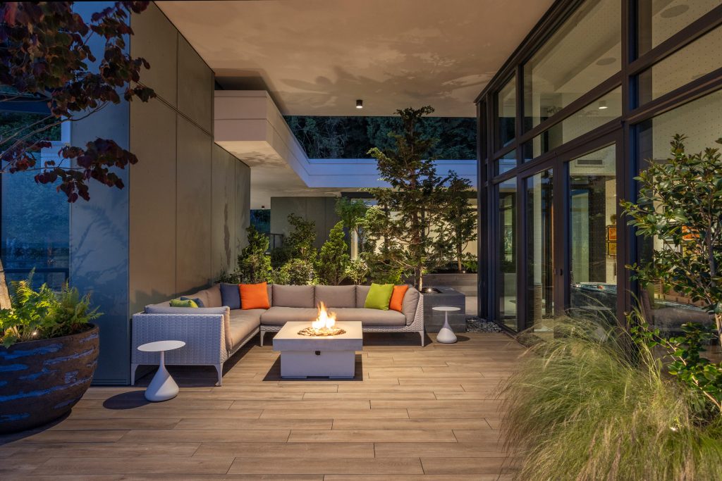 Swicks_3101_Burfield_Web-22-7125 - Swick's Landscaping Ltd. Outdoor patio with an L-shaped sofa and colorful cushions around a fire pit, potted plants, and glass walls under a covered ceiling in evening light.