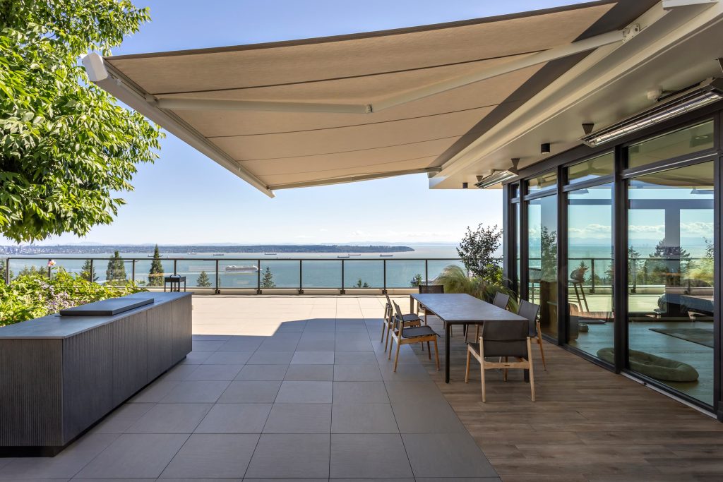 Swicks_3101_Burfield_Web-3-6937 - Swick's Landscaping Ltd. Outdoor terrace with a beige retractable awning, glass railing, and sea view; tables and chairs along the glass wall.