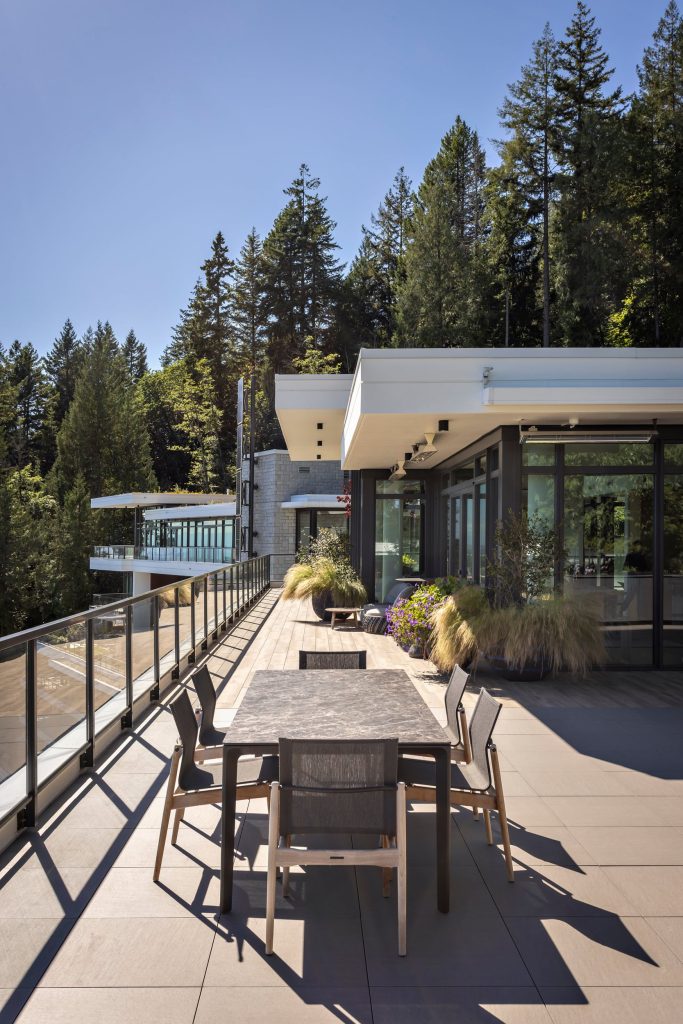 Swicks_3101_Burfield_Web-4-6956 - Swick's Landscaping Ltd. Modern outdoor terrace with a long dining table and chairs, glass railing, and planters, overlooking a forested backdrop under a blue sky.