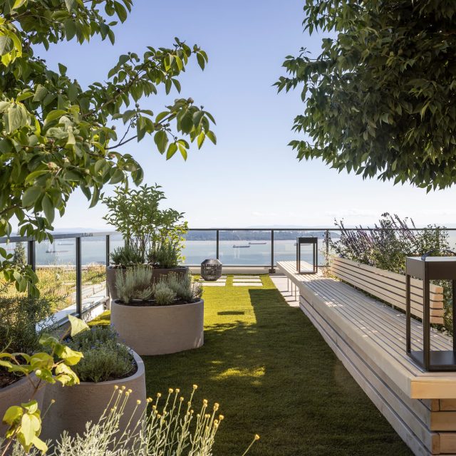 Modern terrace garden with potted plants, wooden benches, and a railing overlooking a body of water outside the space on a sunny day.