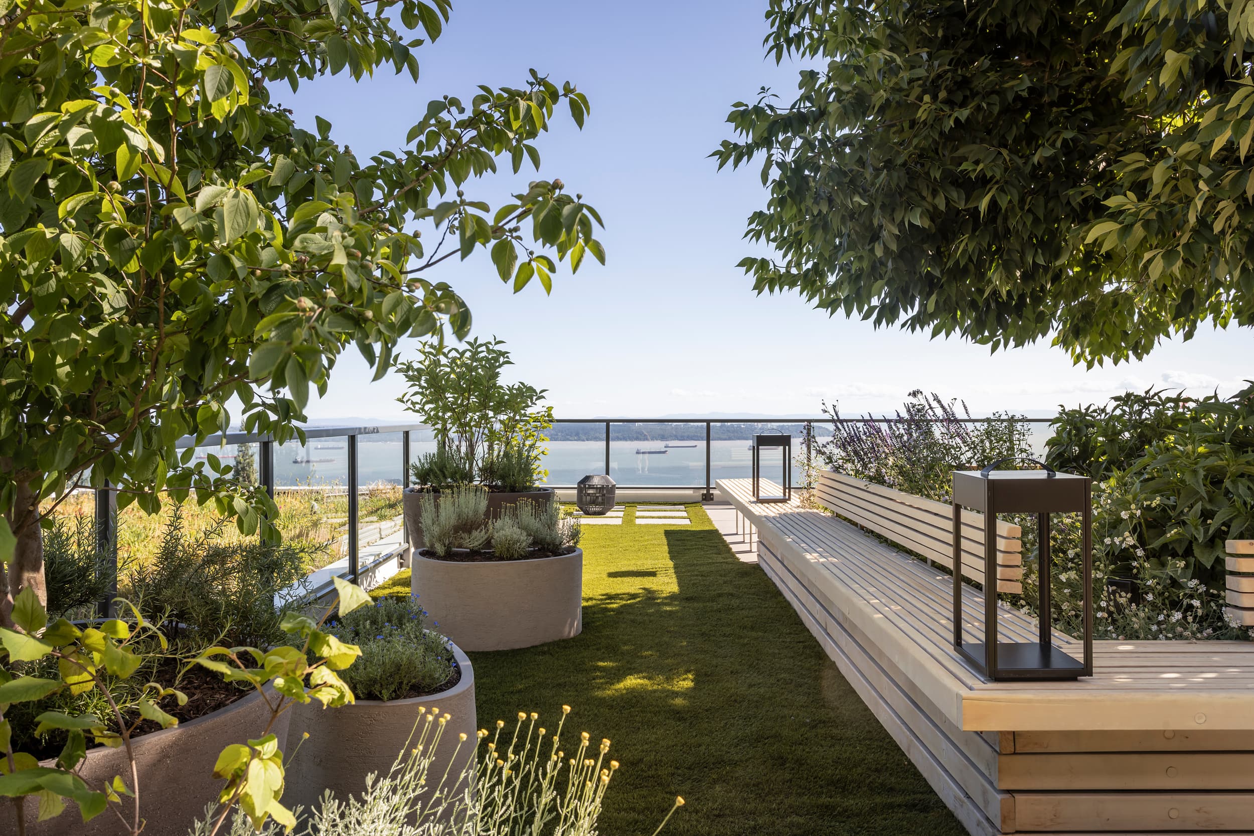 Modern terrace garden with potted plants, wooden benches, and a railing overlooking a body of water outside the space on a sunny day.