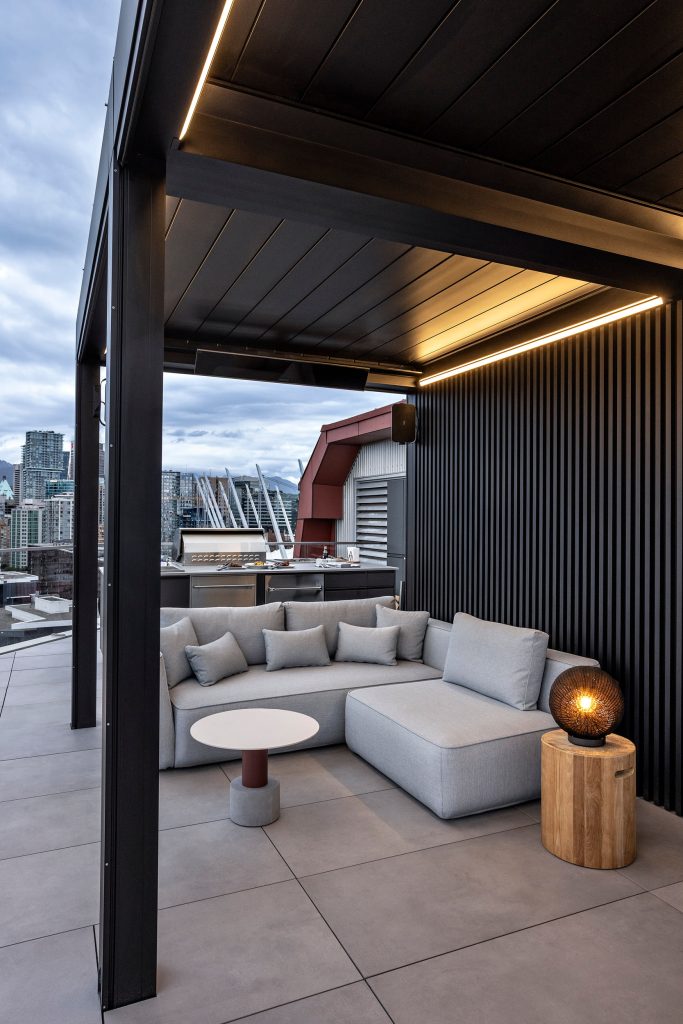 Swicks_918_Cooperage_Web-12-1806 - Swick's Landscaping Ltd. Outdoor rooftop lounge with a gray sectional sofa, round white table, and a warm lantern on a wooden stump, city skyline in the distance.