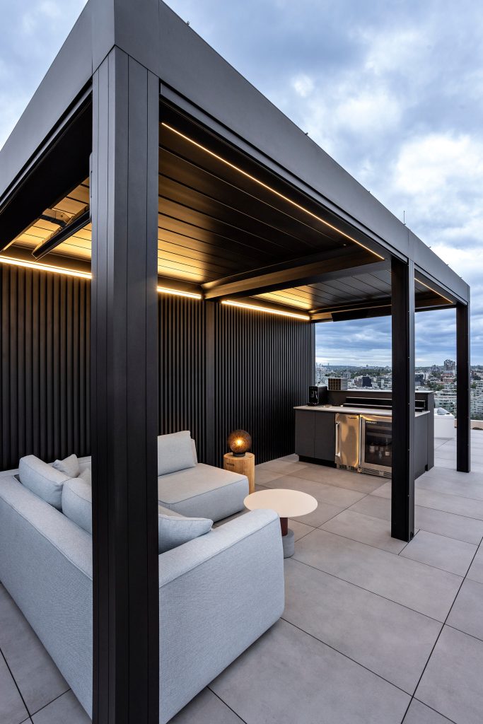 Swicks_918_Cooperage_Web-13-1812 - Swick's Landscaping Ltd. Rooftop living space with black metal pergola, warm LED lighting, and light gray outdoor sofa seating. A built-in grill area is visible in the background with a city skyline beyond.