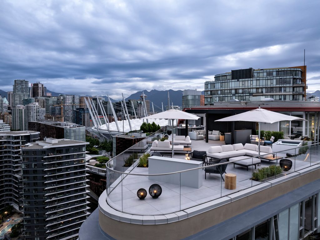 Swicks_918_Cooperage_Web-14-0120 - Swick's Landscaping Ltd. Rooftop lounge with white seating and glass railings, overlooking a city skyline and distant mountains under a cloudy sky.