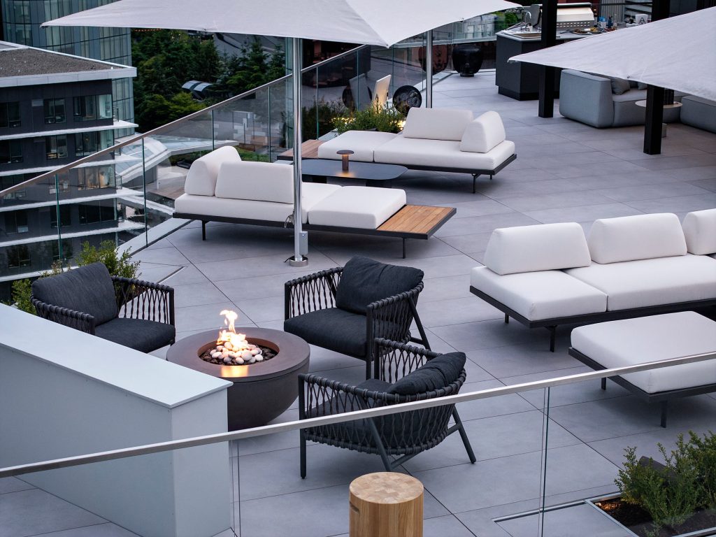 Swicks_918_Cooperage_Web-15-0125 - Swick's Landscaping Ltd. Rooftop lounge with white sectional seating, black wicker chairs, and a lit fire pit under large white umbrellas on a glass-walled terrace.