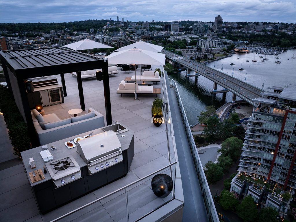 Swicks_918_Cooperage_Web-17-0174 - Swick's Landscaping Ltd. Rooftop outdoor lounge with a built-in grill, white sofas, tables, and large white umbrellas overlooking a city and river.