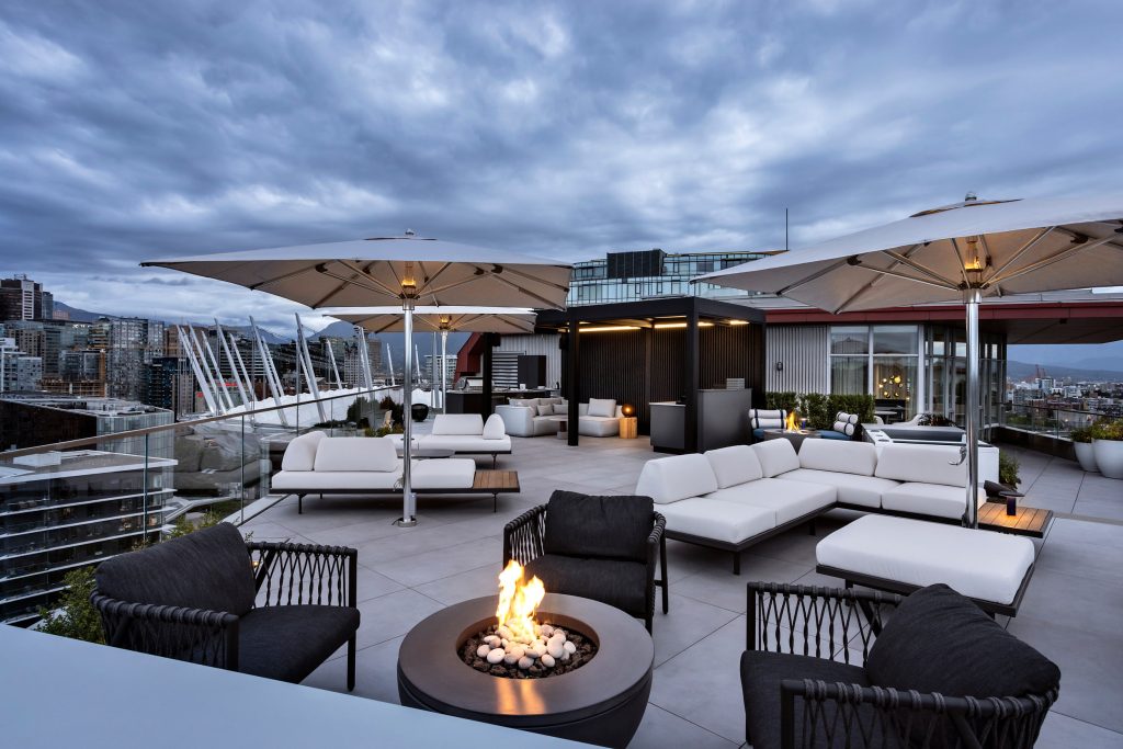 Swicks_918_Cooperage_Web-18-1820 - Swick's Landscaping Ltd. Rooftop lounge with white cushioned seating, large umbrellas, and a fire pit overlooking a cityscape under a cloudy sky.