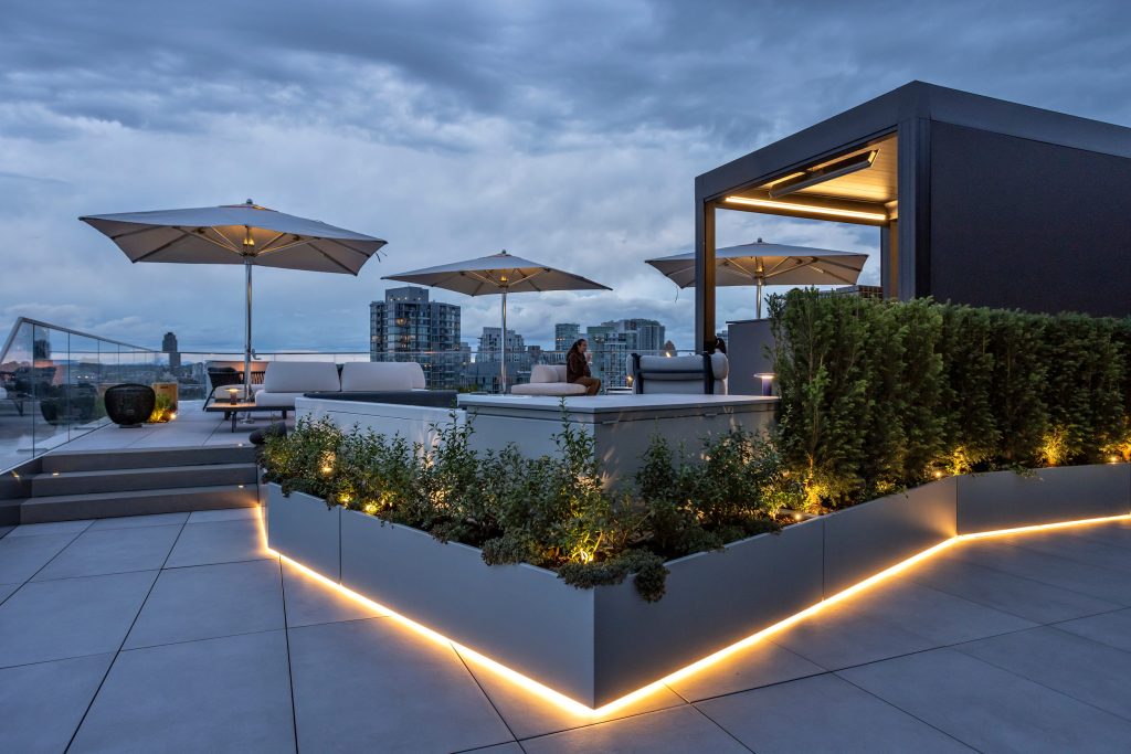 Swicks_918_Cooperage_Web-20-1867 - Swick's Landscaping Ltd. Rooftop lounge area at dusk with white sofas, large umbrellas, glass railing, and warm LED edge lighting; city skyline in the distance under a cloudy sky.