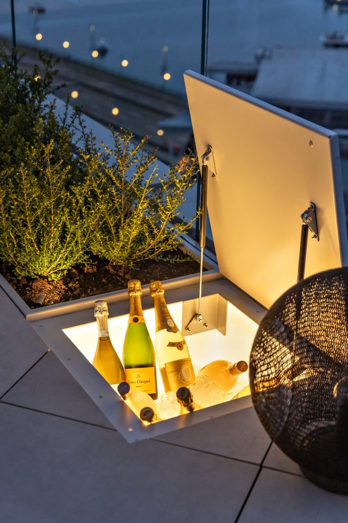 Swicks_918_Cooperage_Web-22-1897 - Swick's Landscaping Ltd. Champagne bottles chilling on ice in an open-lit outdoor bar cooler, with green plants and a woven chair nearby at dusk.