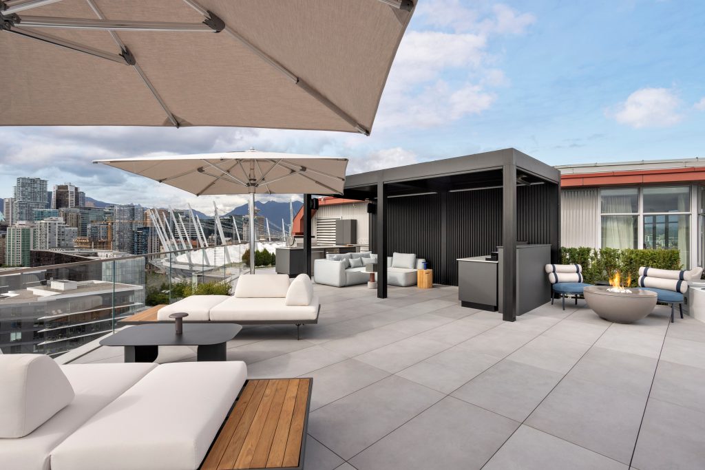 Swicks_918_Cooperage_Web-3-1693 - Swick's Landscaping Ltd. Rooftop terrace with cream sofas, wooden tables, and beige shade sails overlooking a city skyline.