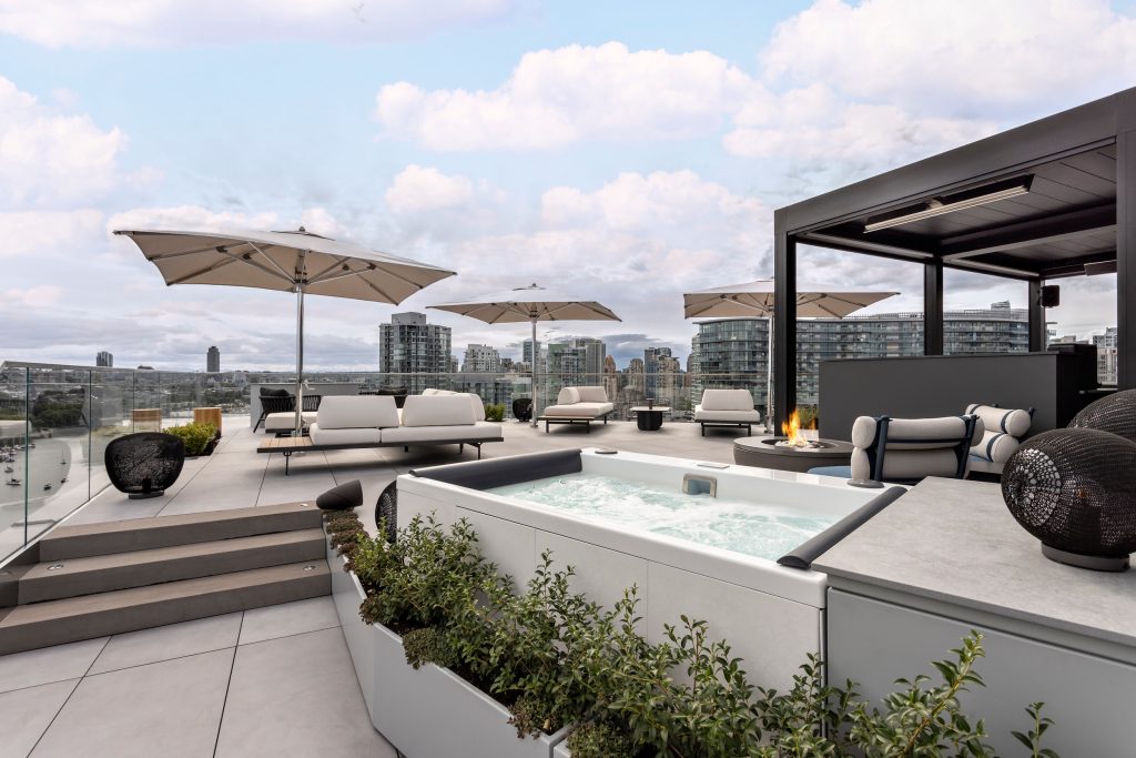 Swicks_918_Cooperage_Web-4-1697 - Swick's Landscaping Ltd. Rooftop lounge with white cushioned seating, large umbrellas, and a hot tub overlooking a city skyline.