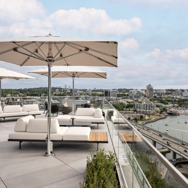 Rooftop outdoor lounge with white sectional couches under large beige umbrellas, overlooking a city harbor.