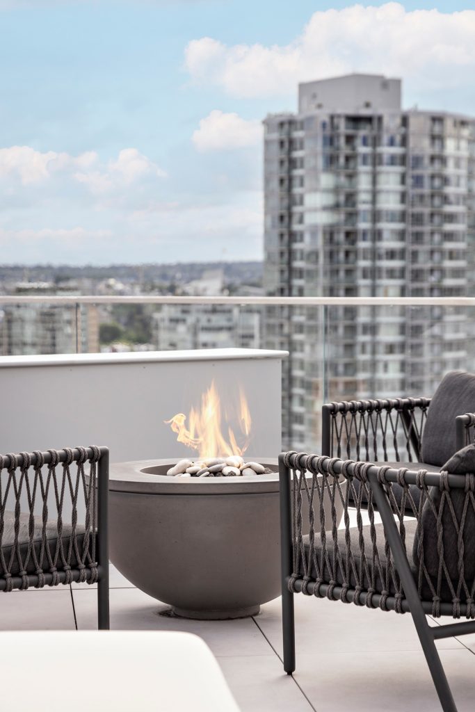 Swicks_918_Cooperage_Web-9-1768 - Swick's Landscaping Ltd. Rooftop patio with a lit fire pit surrounded by woven black chairs and a glass railing, city buildings in the background.