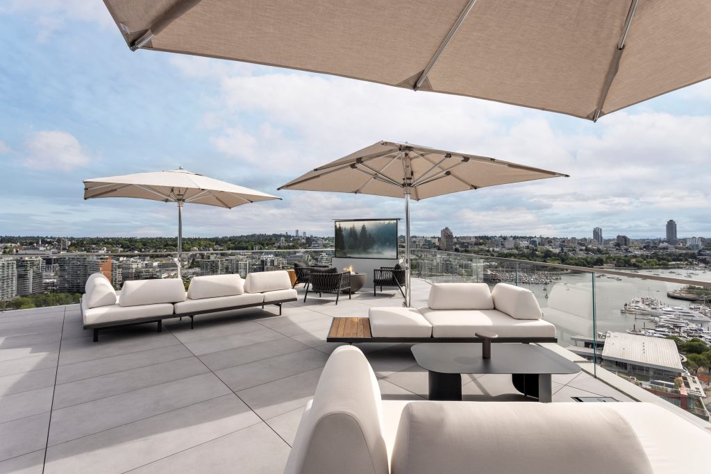 landscape penthouse - Swick's Landscaping Ltd. Rooftop lounge with white sectional seating, beige shade umbrellas, and glass railing overlooking a city marina and skyline.