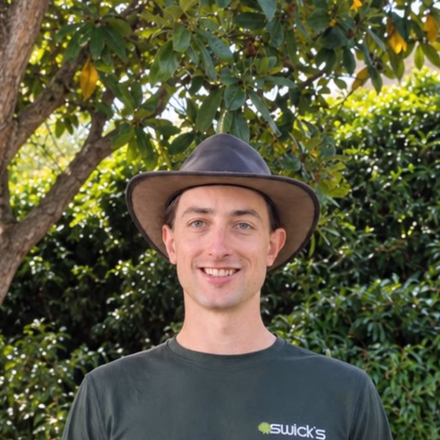 Young man in a wide-brimmed hat and dark green shirt standing outdoors with lush greenery in the background.