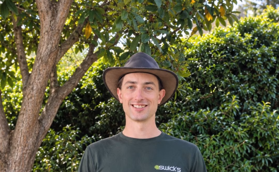 Young man in a wide-brimmed hat and dark green shirt standing outdoors with lush greenery in the background.