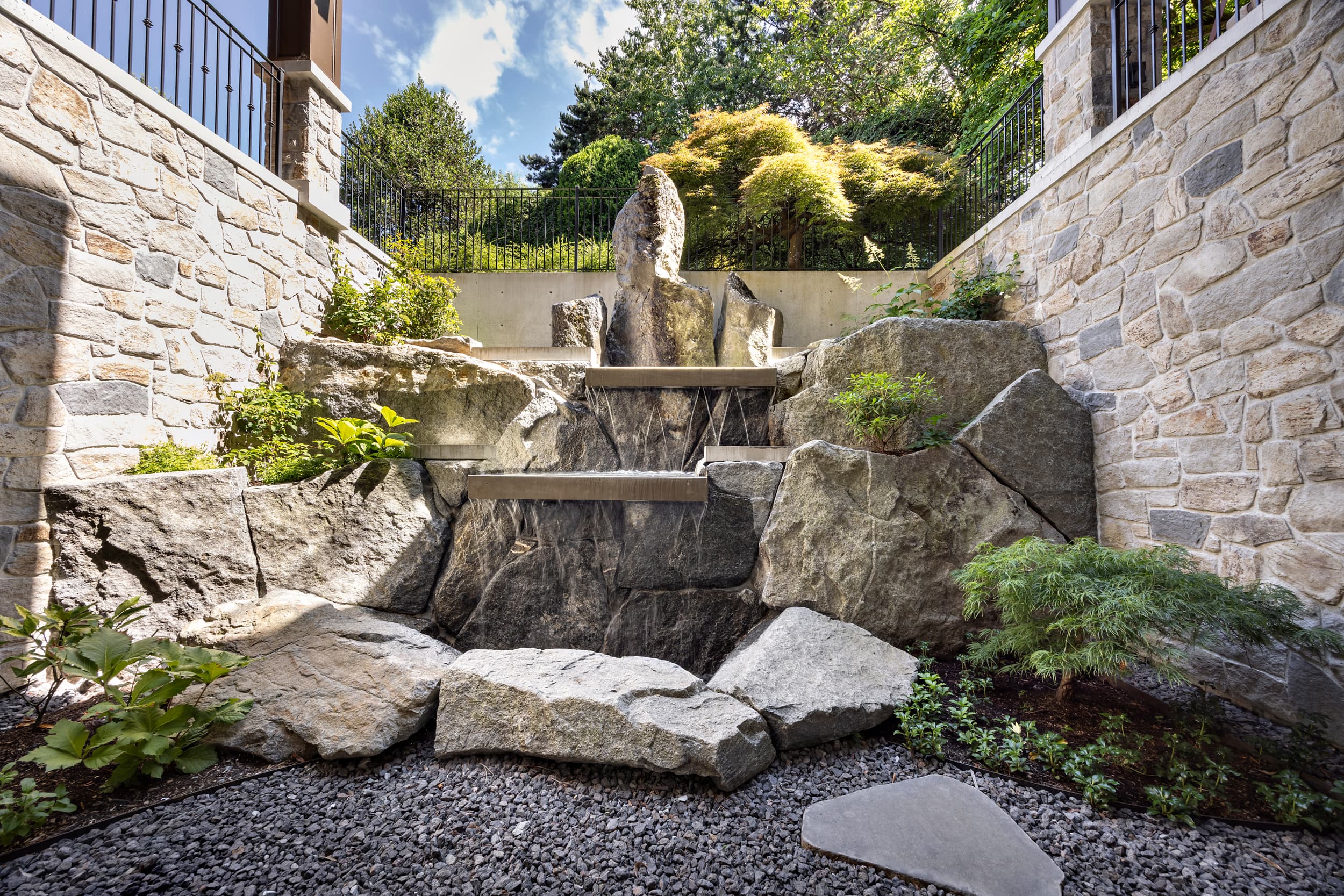 Tiered stone garden with large boulders, shrubs, and greenery between stone retaining walls.