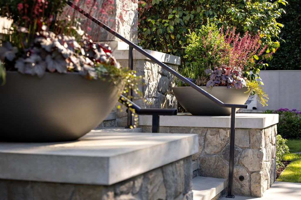 Stone staircase with large round planters and a metal handrail, surrounded by shrubs and flowers.