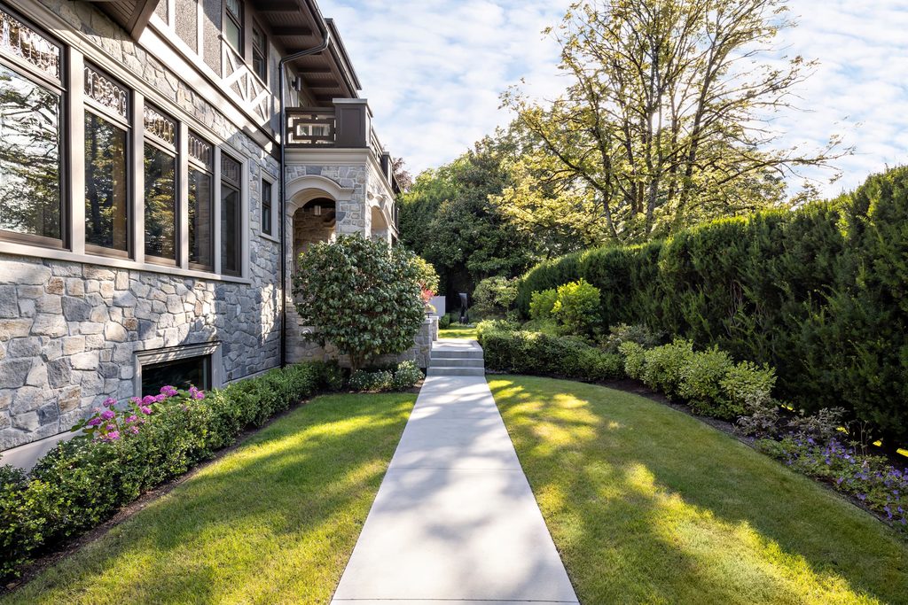 Stone house exterior with large windows, manicured lawn, and a straight concrete walkway by the garden.