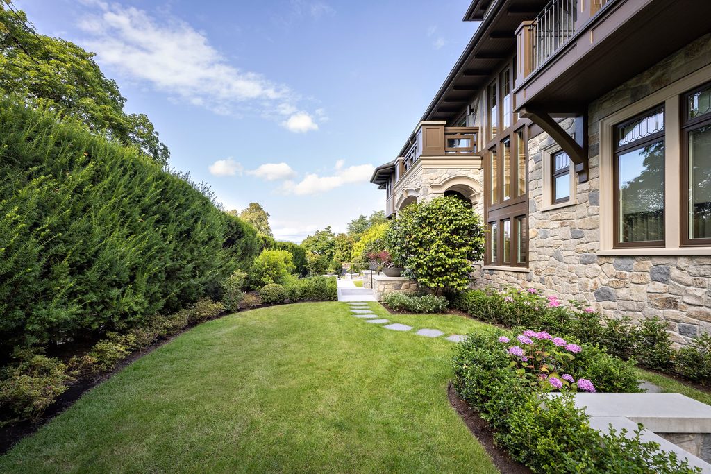 Stone house with large windows and a wooden balcony along a landscaped front yard and stepping-stone path on a sunny day.