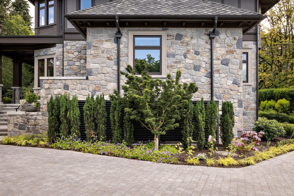 Stone masonry house exterior with beige-framed windows and neatly landscaped front garden along a paved driveway.