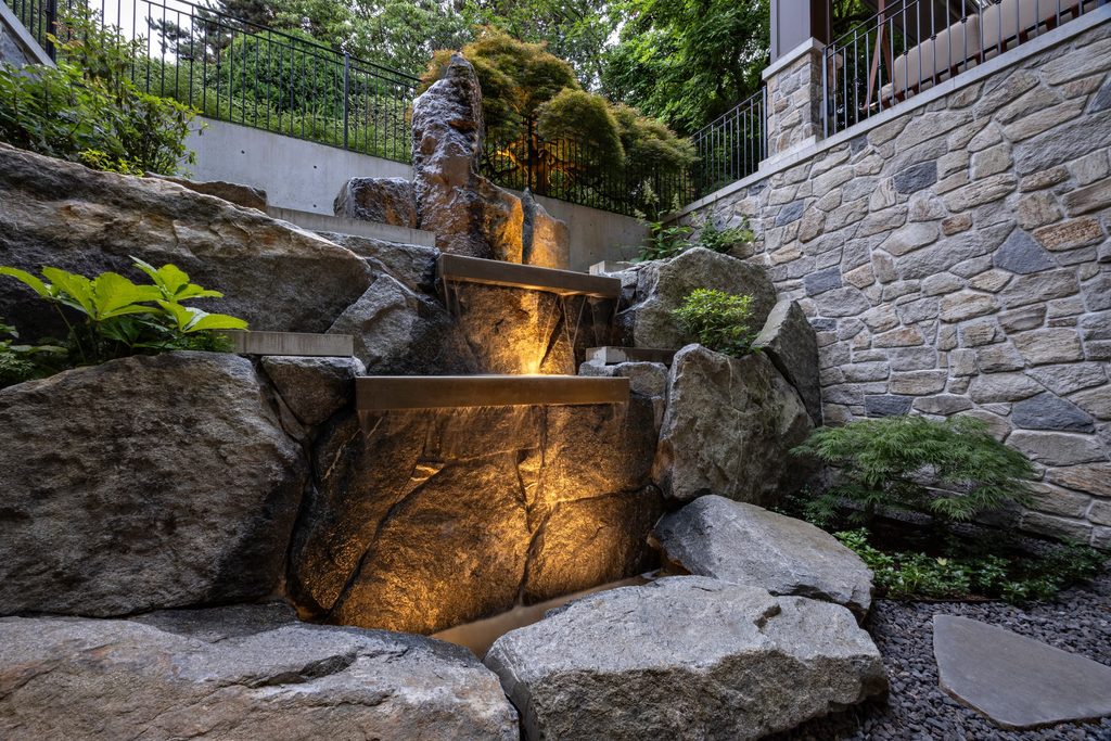 Stone staircase and water feature built from large rocks, illuminated by warm lights, with stone walls and greenery in a landscaped garden.