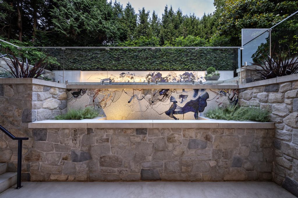 Outdoor stone terrace with a glass railing above a mosaic wall depicting people dancing; dense greenery in the background behind the railing.