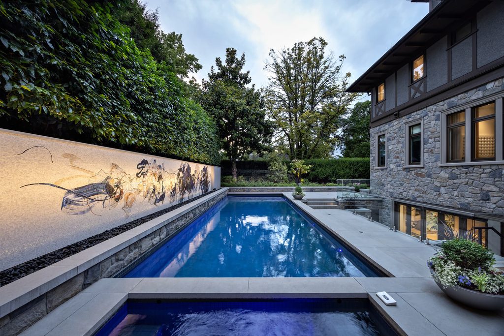 Outdoor pool in a courtyard beside a stone house, with a mural wall on the left, hedges and trees behind, and lit interior windows.