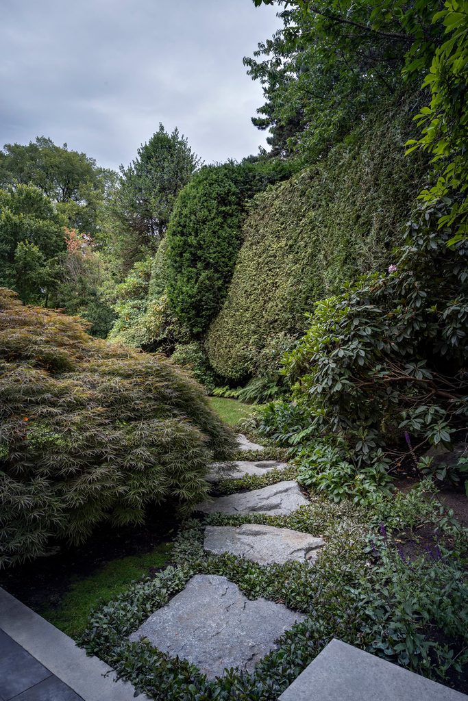 Stone stepping path winding through a dense, manicured garden with tall hedges and trees overhead on a cloudy day.