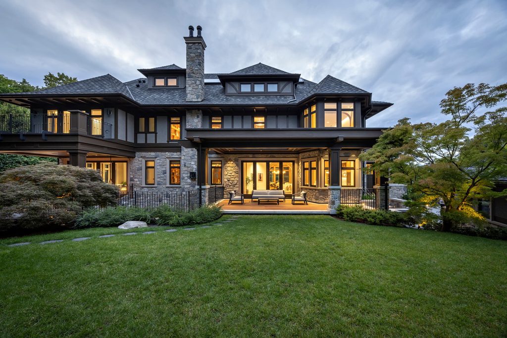 Modern two‑story stone and dark wood house with lit windows and a covered patio seating area, set on a green lawn at dusk.