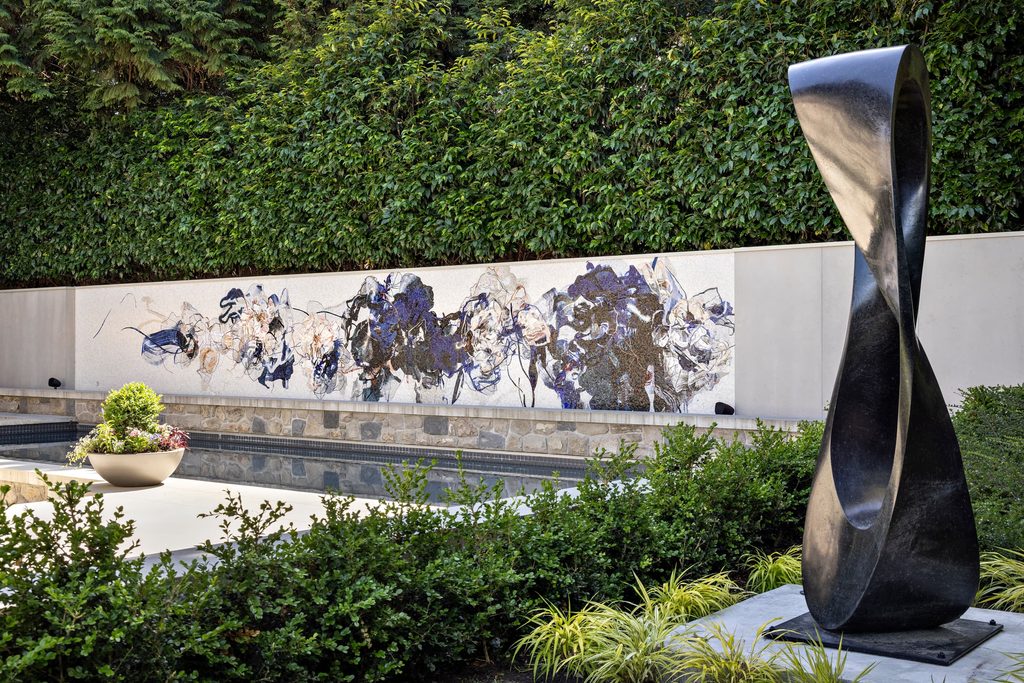 Abstract outdoor sculpture beside a twisting black metal form, with a mural on a low wall and a dense hedge behind.