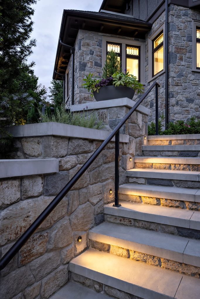 Stone house exterior with a stone staircase and black metal railing, leading to a warmly lit doorway and windows; a large planter with greenery sits on a ledge above the stairs.