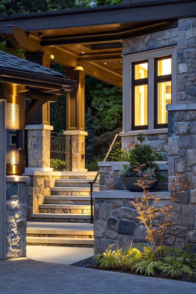 Stone home's entryway at dusk with warm window light, stone walls, and timber columns along illuminated stone stairs surrounded by greenery.