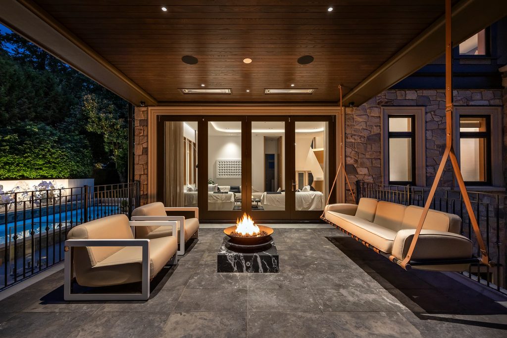 Outdoor lounge area at night with beige cushioned chairs around a fire pit on a dark tiled patio under a wood ceiling. ‏‏(ambience shot)