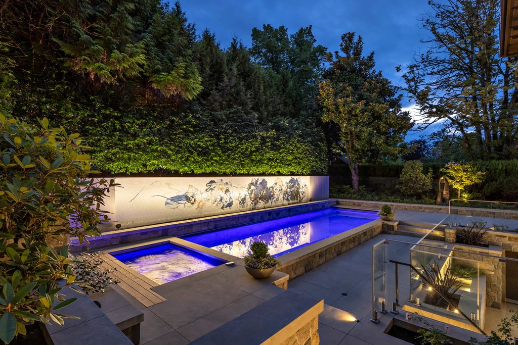Evening backyard with a blue-lit rectangular pool, glass railing, and a long mural wall surrounded by trees.