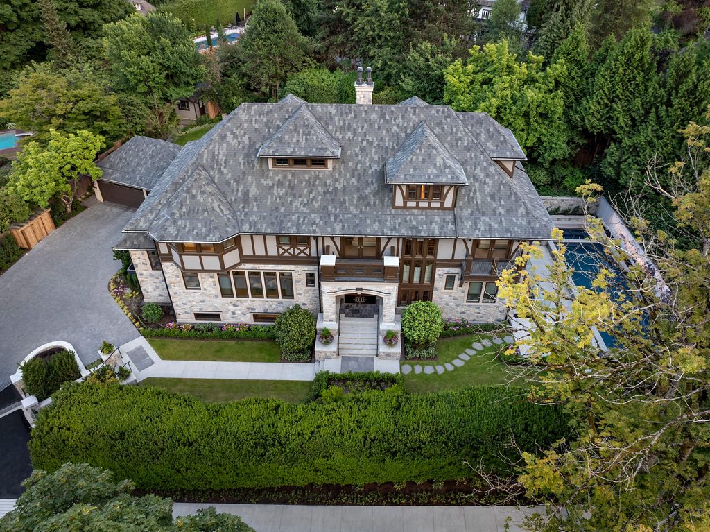Aerial view of a large Tudor-style mansion with a gray shingled roof, stone facade, and surrounding green trees and manicured lawn nearby.