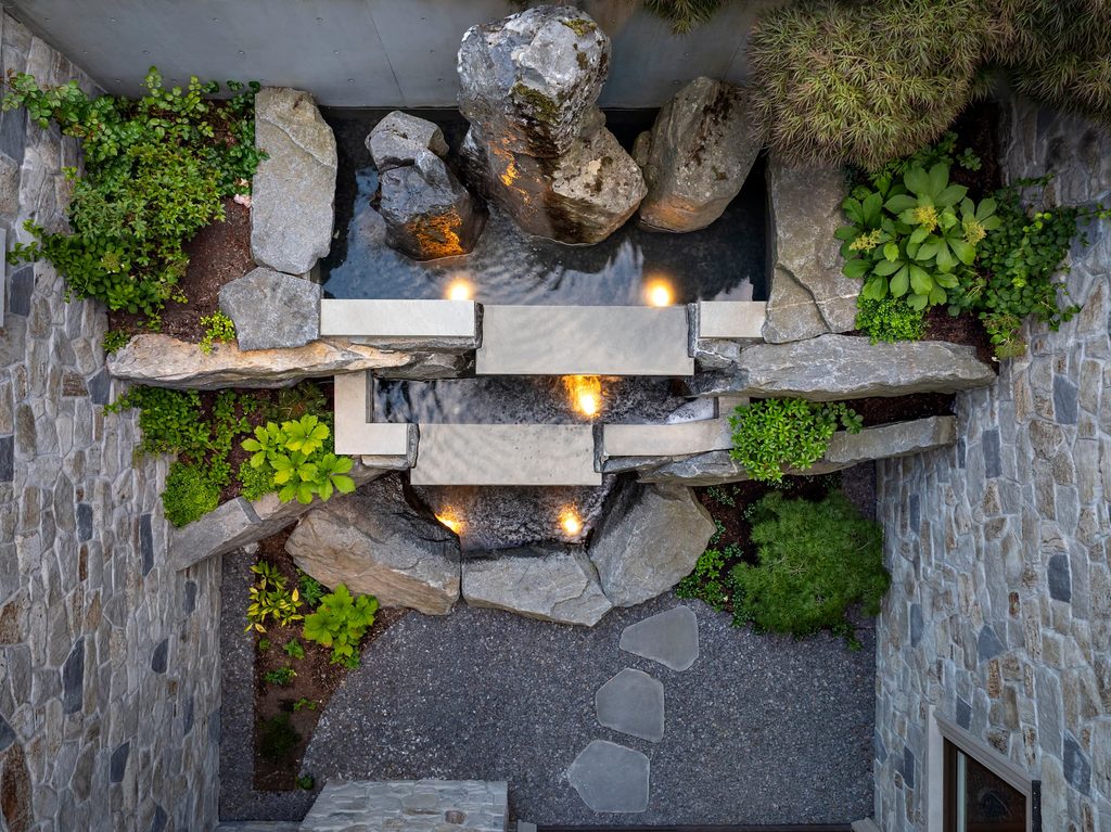 Top-down view of a stone courtyard featuring a rectangular water basin with glowing lights, large rocks, stepping stones, and surrounding greenery.