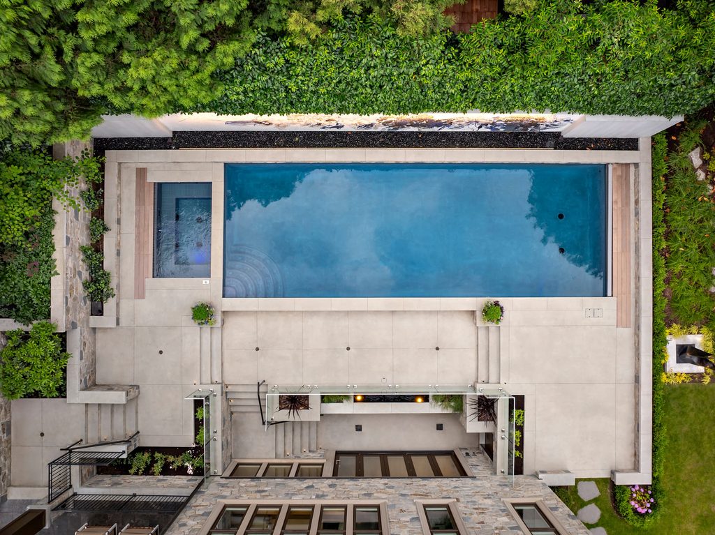 Aerial view of a rectangular blue pool surrounded by light gray concrete and lush green hedges along the top edge.