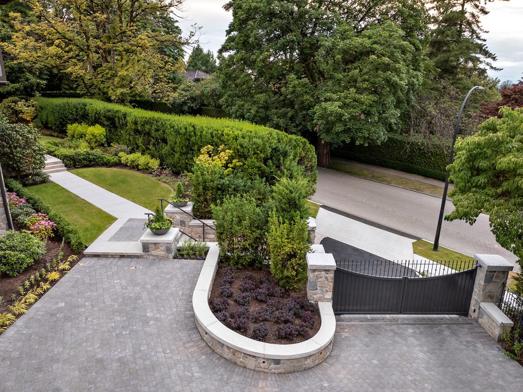 Circular stone planter with purple foliage at a paved driveway and front steps, greenery and hedges surrounding a modern gate.