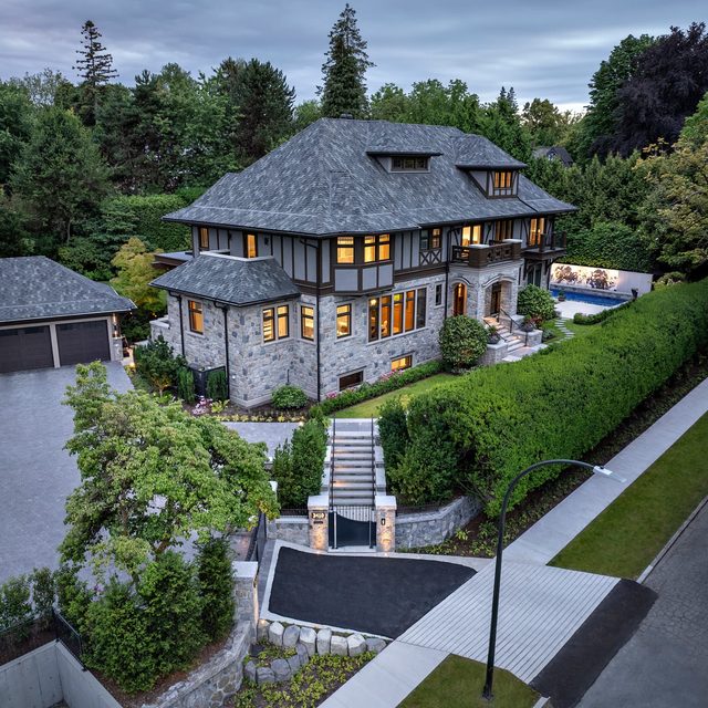 A large upscale stone-and-wood mansion with a slate roof, viewed from above among tall trees and a manicured hedge.