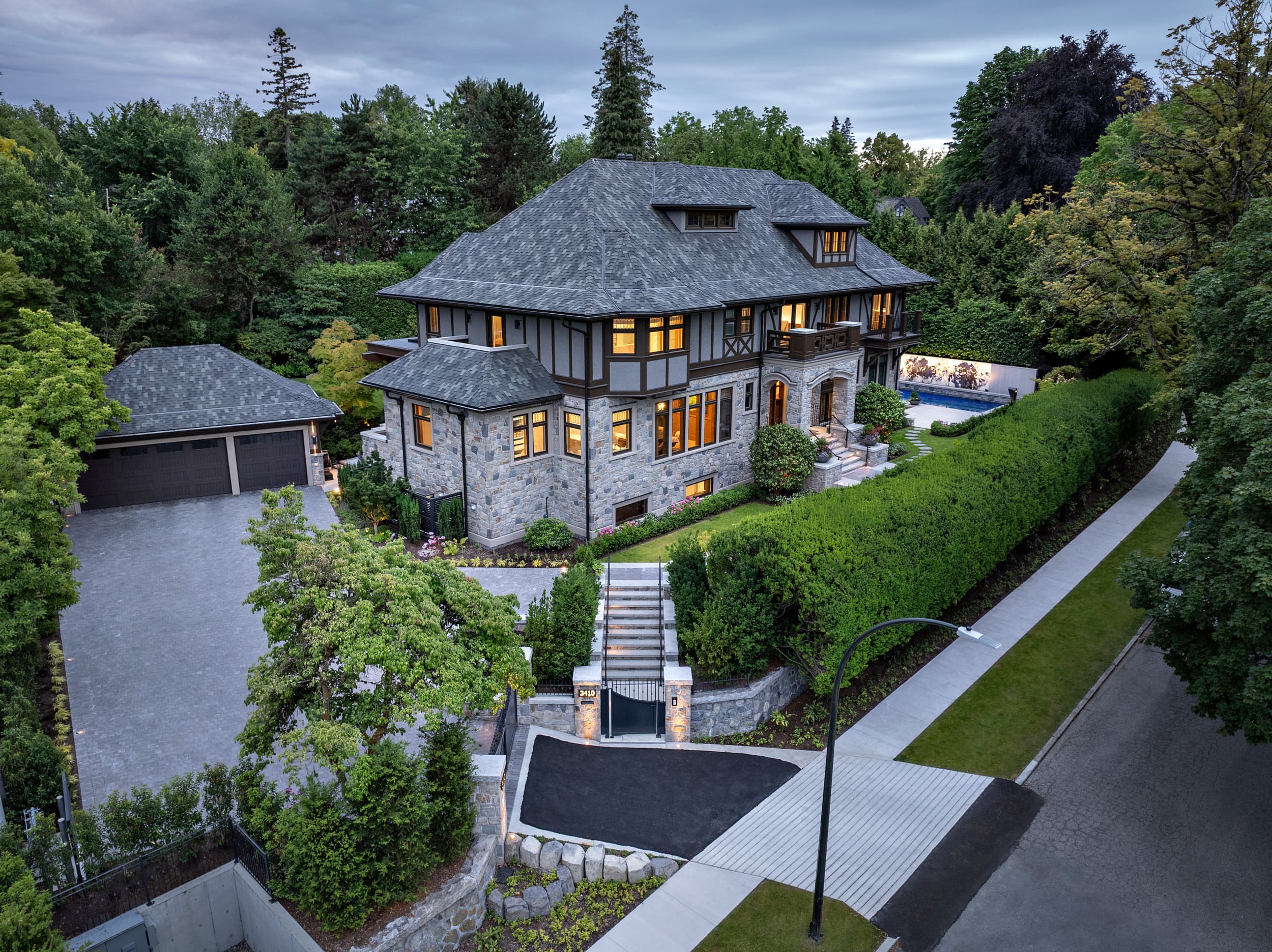 A large upscale stone-and-wood mansion with a slate roof, viewed from above among tall trees and a manicured hedge.