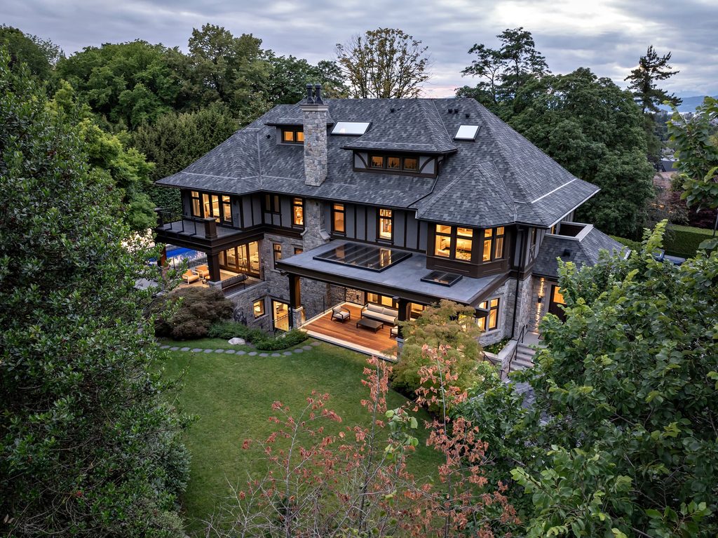 Luxurious house with dark slate roof, stone chimney, and large windows among trees in a lush yard.
