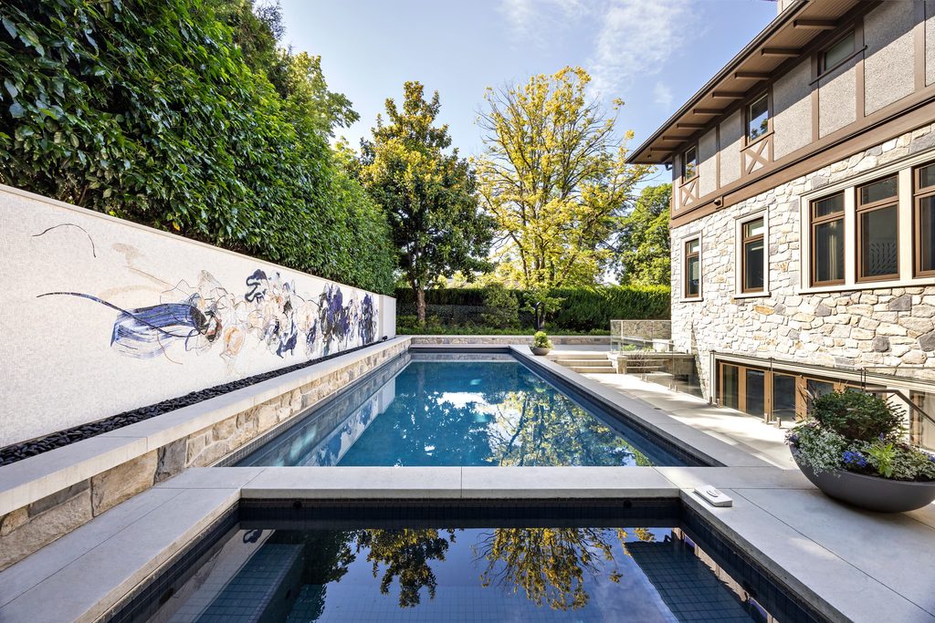 Rectangular outdoor pool beside a stone house, with hedges, trees, and a mural wall on the left.