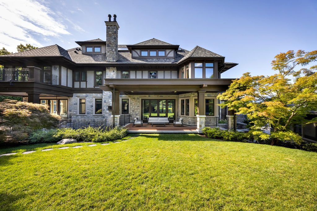 Luxury stone-and-wood house with a covered patio and outdoor seating on a sunny lawn.
